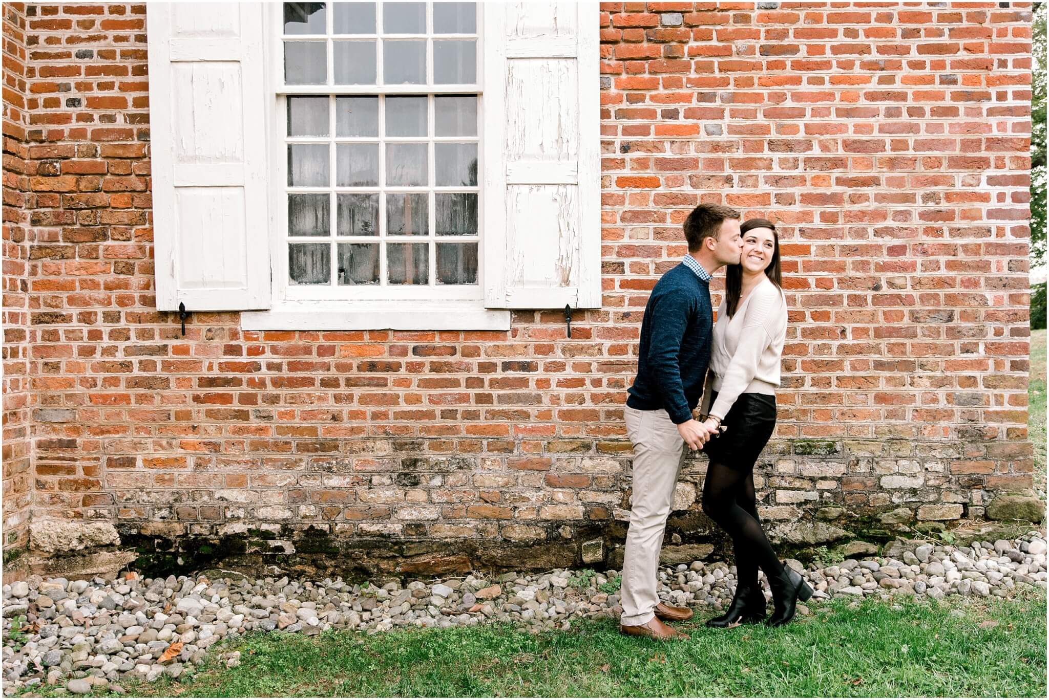 man kissing fiance on the cheek in front of a brick wall in annapolis 