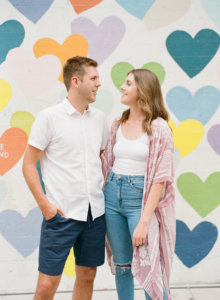 Couple in front of multi colored heart wall looking at each other and smiling